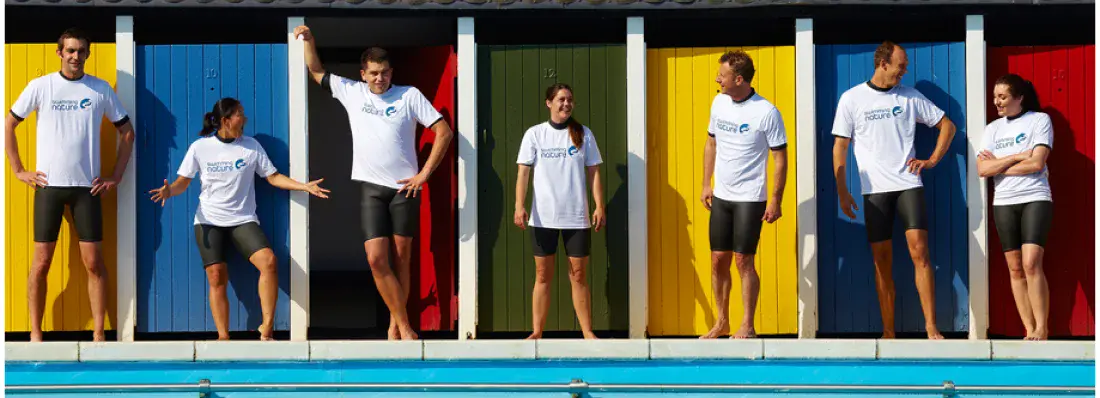 Eight Swimming Nature instructors pose playfully in front of a row of brightly coloured beach‑hut doors, each raising an arm in celebration.