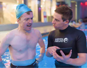 Smiling adult swimmer in a blue cap chats poolside with a Swimming Nature instructor in a black rash‑vest.