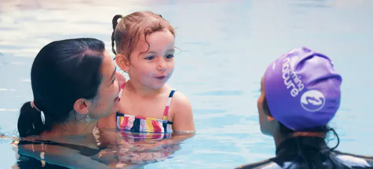Little girl in a colourful striped swimsuit listens attentively to an instructor while her mum supports her chest‑deep in the pool.