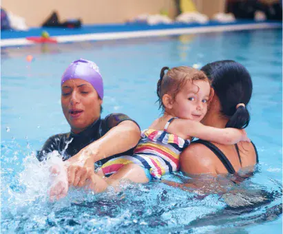 Instructor guides a toddler’s splashing kick as the child clings to mum’s shoulder in the water.