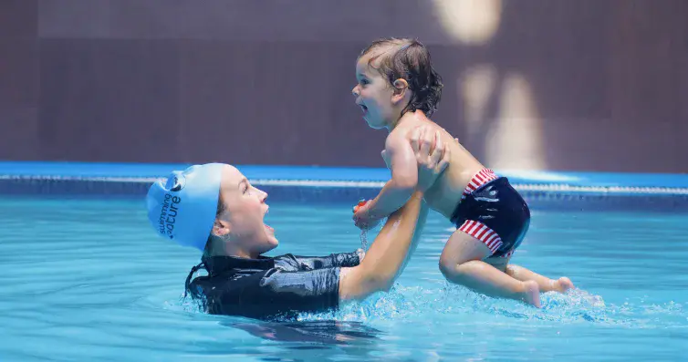 Swimming Nature instructor in blue cap joyfully lifts a laughing toddler from the water in a baby‑and‑parent swim class.