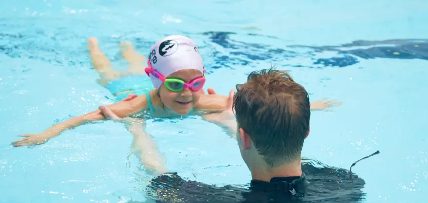 Swimming instructor in the pool guides a young girl wearing pink goggles and a white swim cap as she glides face‑down across the water.