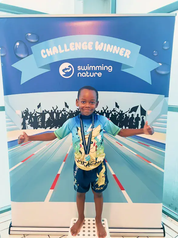 Smiling boy wearing multiple medals spreads his arms in front of a “Challenge Winner” backdrop beside a pool lane graphic.