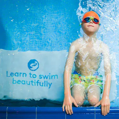 Child in orange goggles and swim cap stands under a cascade of water beside a “Learn to swim beautifully” banner.
