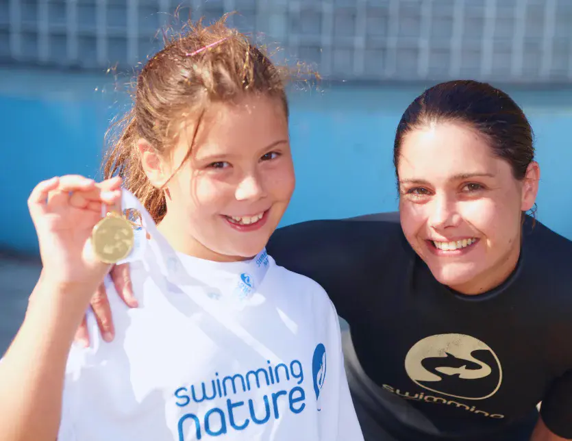 Young girl proudly shows a gold swimming medal while her instructor kneels beside her; both grin at the camera poolside.
