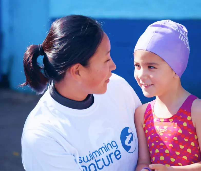 Swimming Nature instructor in a branded T‑shirt chats warmly with a girl in a pink cap and spotty swimsuit at the poolside.