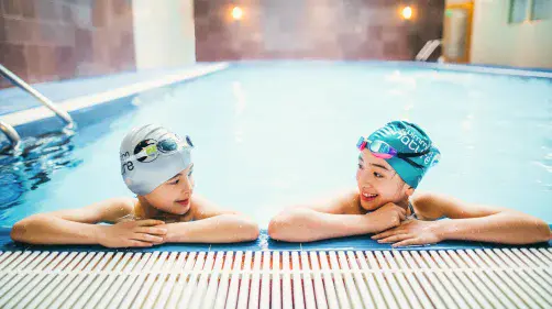 Two swimmers in blue and white caps relax at the pool edge, chatting shoulder‑to‑shoulder in calm turquoise water.