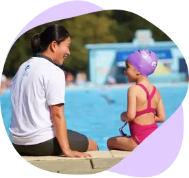 Swimming Nature instructor in a white polo shirt chats with a girl in a purple cap and pink swimsuit seated on the pool edge.