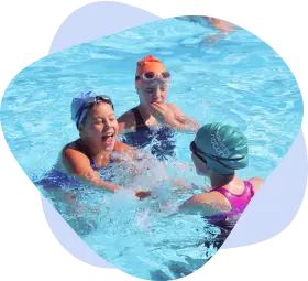 Two primary‑age pupils in colourful caps laugh and splash water at each other during a fun pool session.
