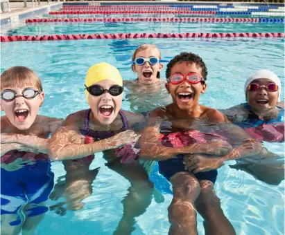 Group of laughing children in goggles and caps cling to the lane rope, posing together in the pool.