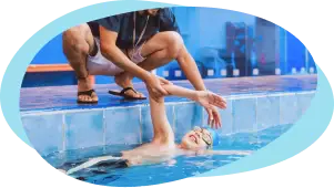 Coach kneels pool‑side helping a child practise front‑crawl kicks at the water’s edge.