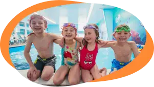 Four smiling youngsters in colourful caps sit arm‑in‑arm on the poolside after class.